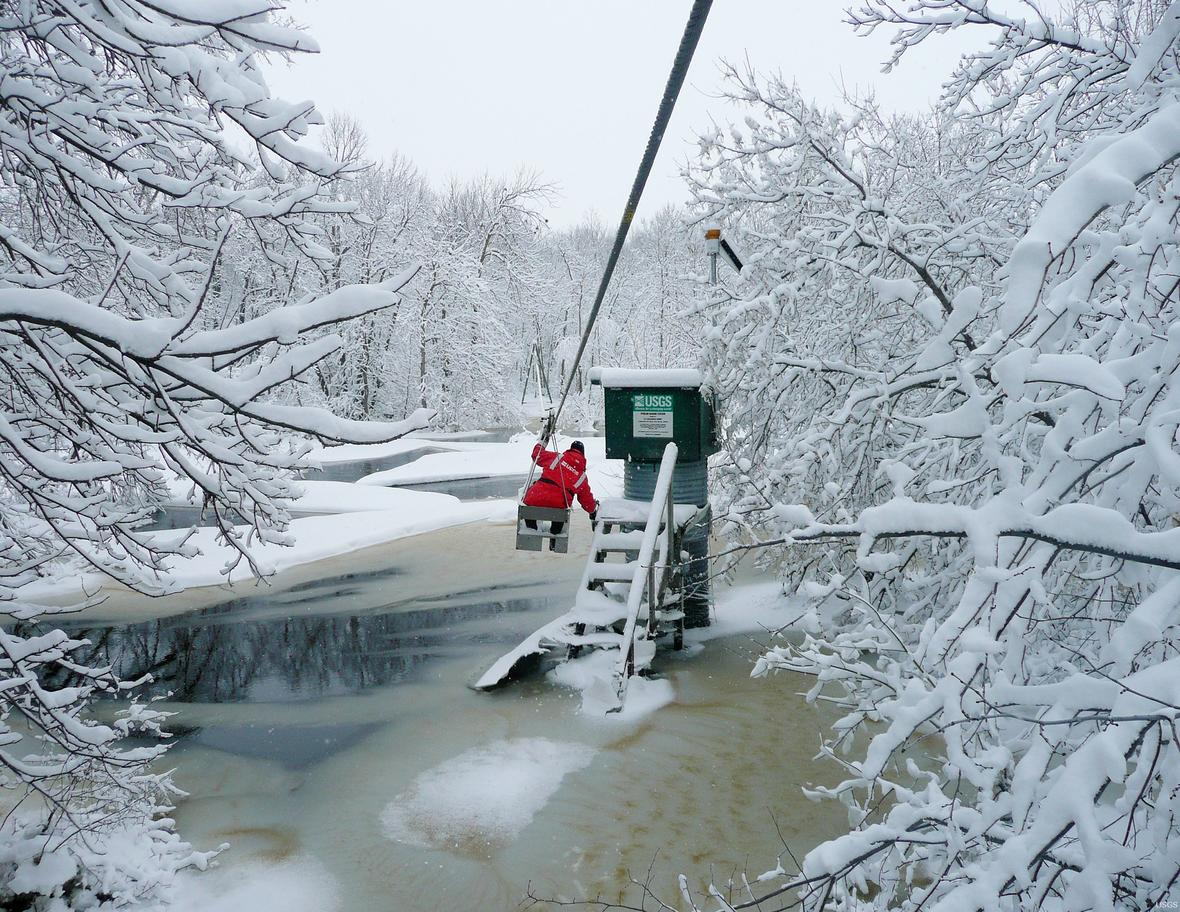 A man hangs from a cable car suspended over a river. He is moving towards a streamgage over the Roseau Rivernear Malun, MN.