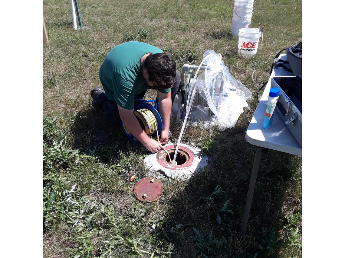 Picture of a person in the grass kneeling over a circular concrete wellhead, with instruments in the background