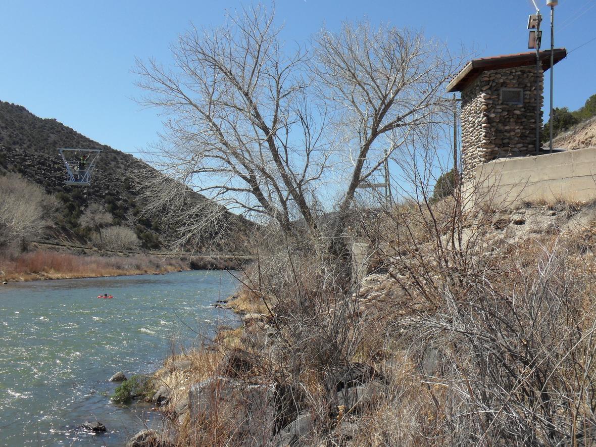 Image of streamgage on the Rio Grande at Embudo. Gage house made of local stone, cableway for measuring flow, riffles, and a person on a kayak in river.