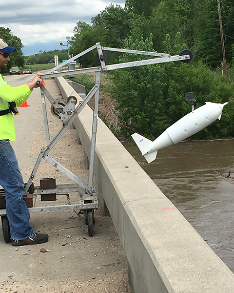 A person standing on a bridge operates a crane with a sediment sampling device.