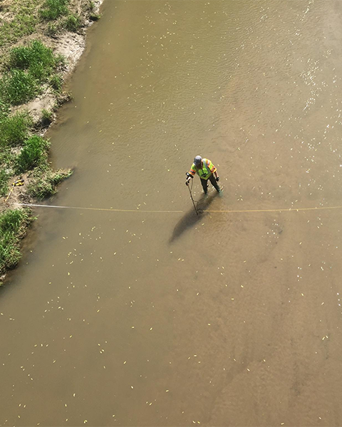 A person stands in a brown river and uses a pole to collect a sample of water